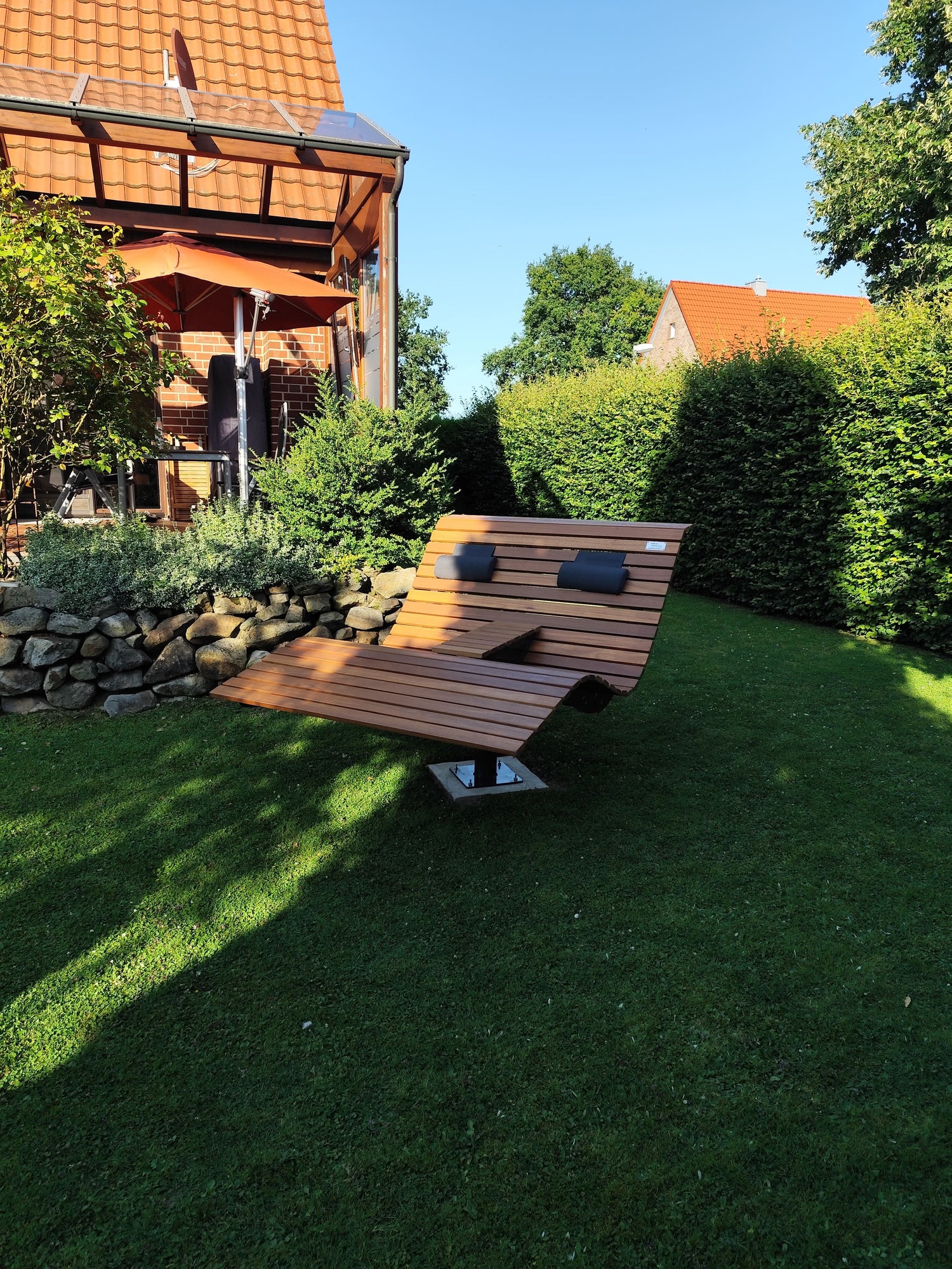 Ein elegantes, drehbares Waldsofa mit Holzlatten und zwei Polsterkissen, aufgestellt auf einer gepflegten Rasenfläche. Im Hintergrund befindet sich ein Haus mit roten Ziegeln und einem Glas-Wintergarten, flankiert von dichten Hecken und Sträuchern. Die Szene ist sonnig, mit klarem blauem Himmel, und vermittelt eine gemütliche Gartenatmosphäre.